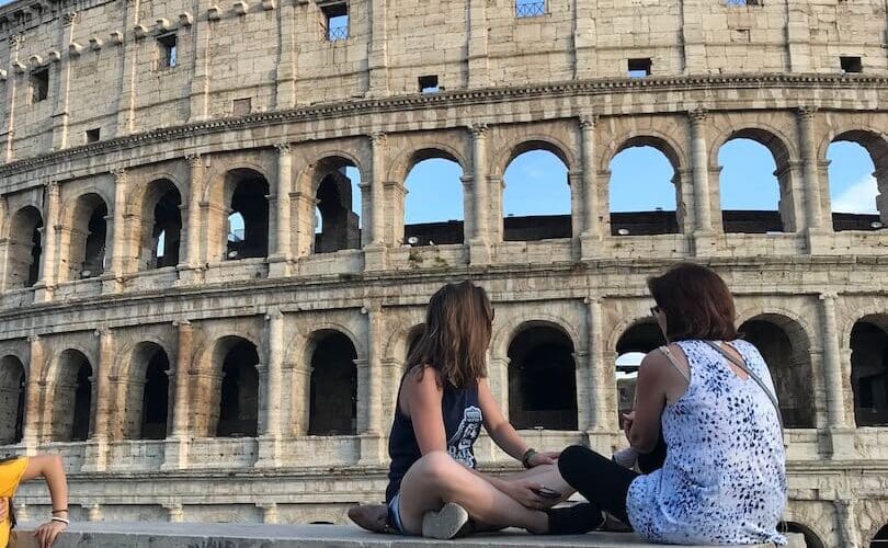 Two women sitting criss cross on a wall in front of the Colosseum in Rome
