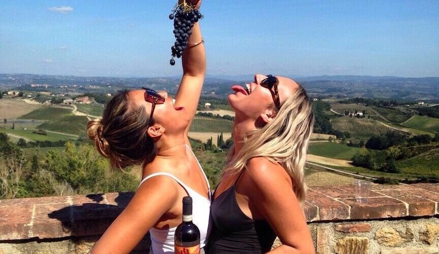 Two laughing friends pretending to eat a bunch of grapes while holding a bottle of wine as well