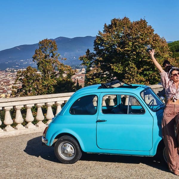 Woman happily leaning against a bright blue fiat 500 and a view on Florence