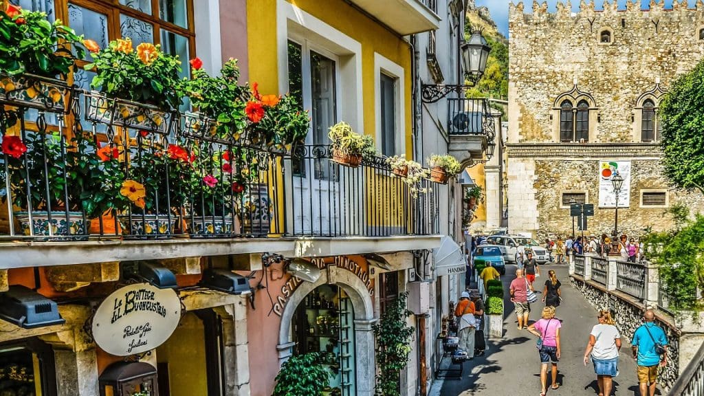 Balcony adorned with flowers overlooking a bustling street of Taormina Sicily near historical buildings.