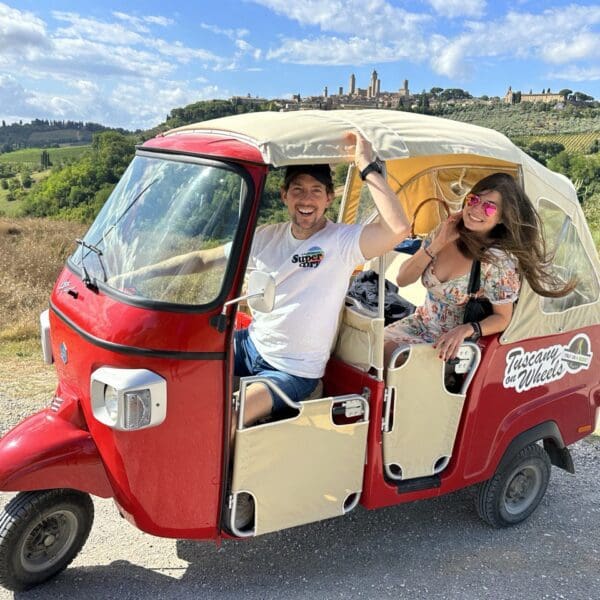 Man in the front seat of a red tuk tuk, and a woman in the back, with San Gimignano in the background